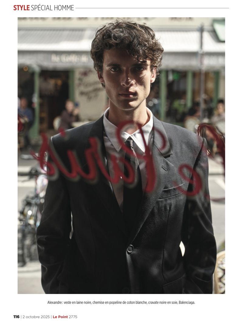 Editorial shot of a man in a black suit and tie with dark curly hair, captured through a glass window with red painted text.