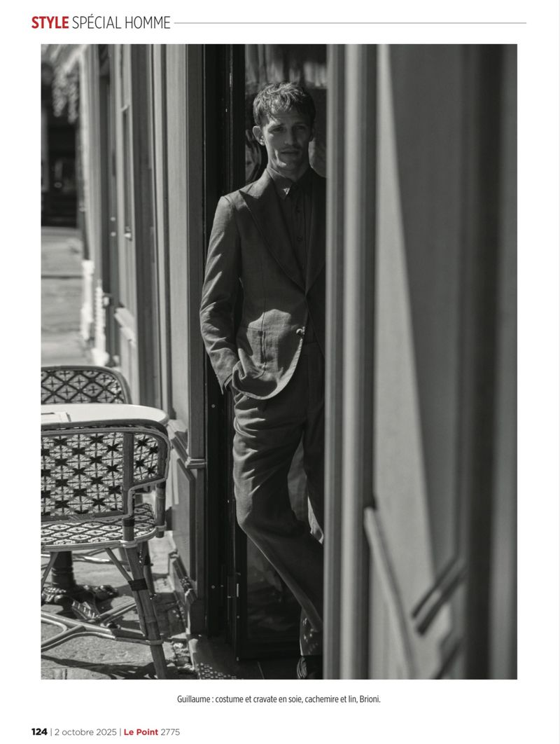 Black and white editorial photograph of a man in a tailored suit standing in a cafe doorway beside woven bistro chairs.