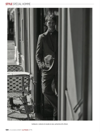 Black and white editorial photograph of a man in a tailored suit standing in a cafe doorway beside woven bistro chairs.
