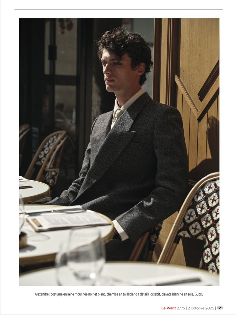 A man in a charcoal grey suit jacket seated at a bistro table outside on a sunny day