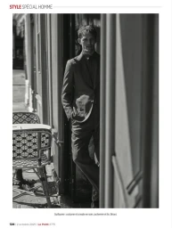 Black and white editorial photograph of a man in a tailored suit standing in a cafe doorway beside woven bistro chairs.