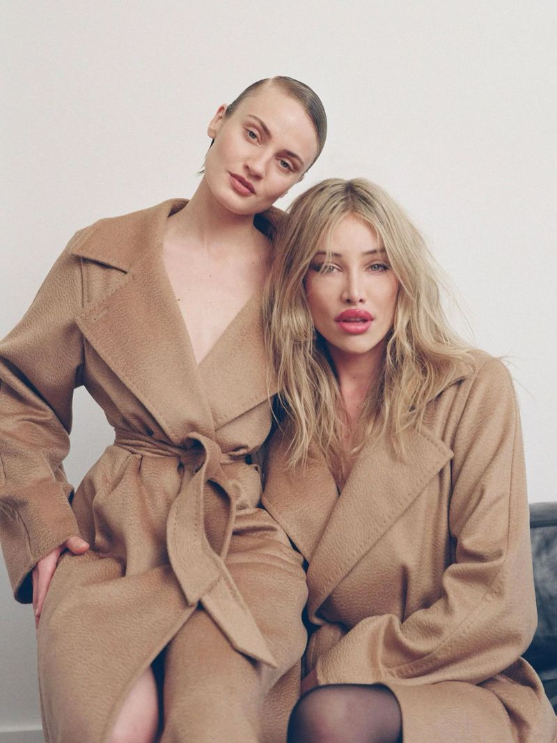 Two blonde fashion models in tan coats pose in front of an off-white wall in a studio. Beauty and fashion editorial photography.
