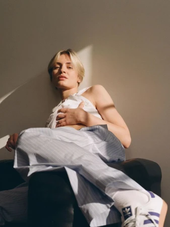 A woman with short blonde hair sits in an armchair wearing a white top and striped pants in a sunlit room, looking at the camera.