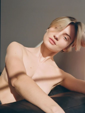 Portrait of a fair-skinned young adult woman with short blonde hair, posing indoors with soft natural light and shadows against a gray wall.