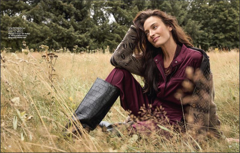 A woman with long brown hair is sitting in a grassy field, wearing a burgundy jumpsuit, dark boots, and a patterned sweater.