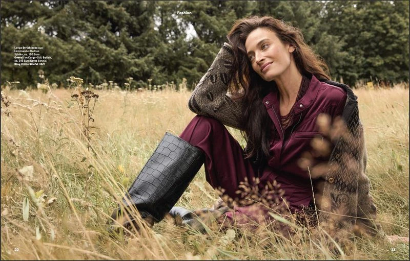 A woman with long brown hair is sitting in a grassy field, wearing a burgundy jumpsuit, dark boots, and a patterned sweater.