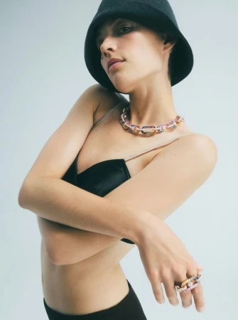 Adult female fashion model in black bra top and hat, wearing chain necklace and ring poses in a medium shot on white backdrop.