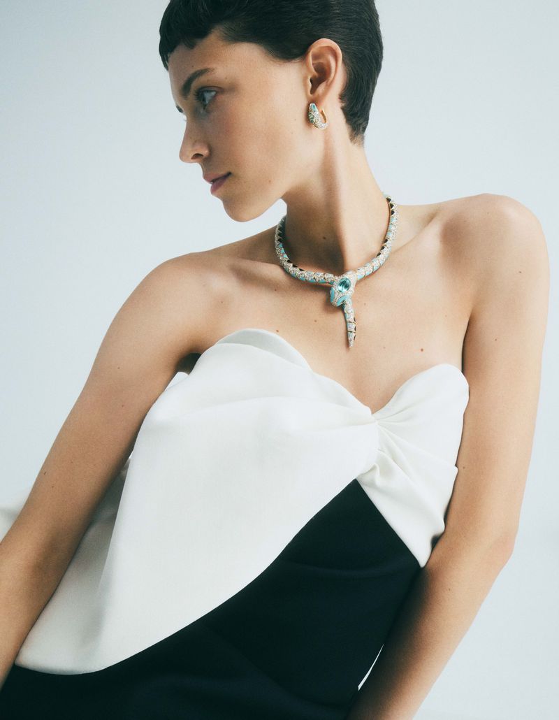 A fashionable woman with a black-and-white dress wearing a diamond and turquoise serpent necklace and earrings against a light backdrop.