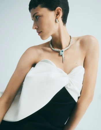 A fashionable woman with a black-and-white dress wearing a diamond and turquoise serpent necklace and earrings against a light backdrop.