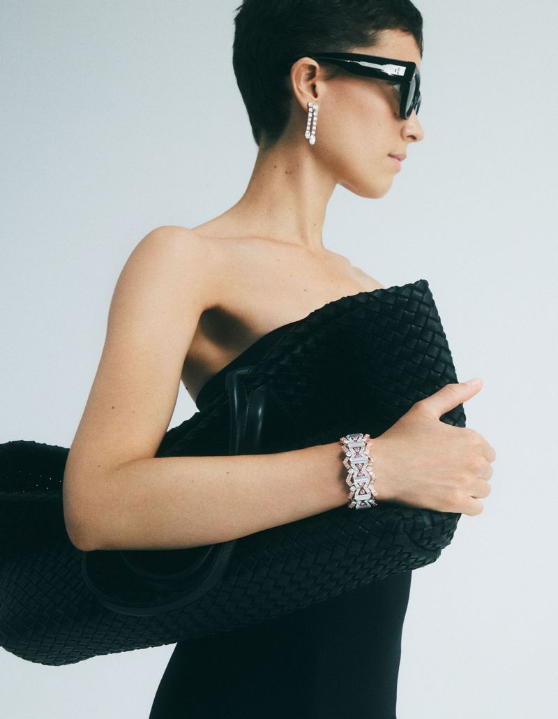 Woman with short hair wearing sunglasses and a black dress, holding a woven black bag and a bracelet against a white backdrop.