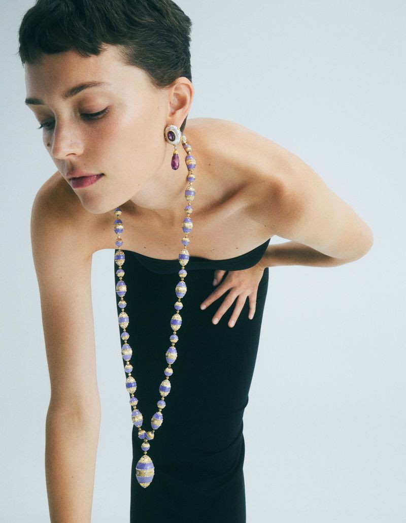 Woman poses in black top with long beaded necklace and matching earrings against a white background in a fashion studio shot.