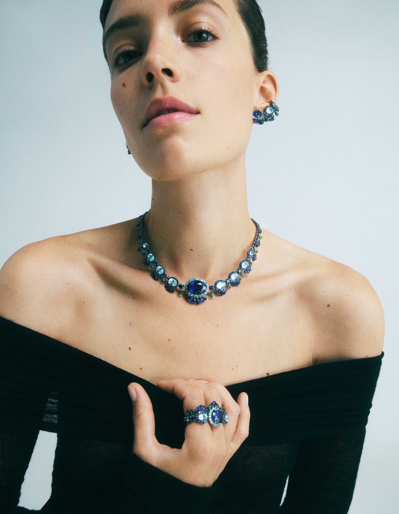 Woman showcases blue gemstone jewelry set featuring a necklace, ring, and earrings, set against a light background in a studio setting.