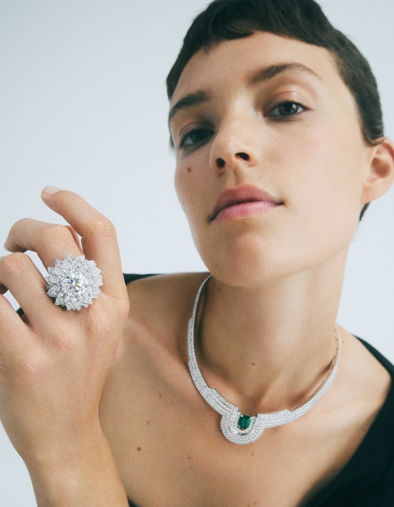 Adult female with pixie hairstyle poses in a studio with silver and green emerald jewelry, including a necklace and ring.
