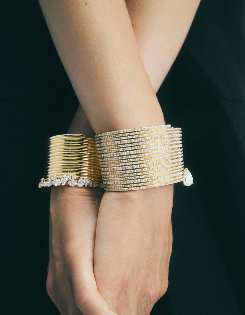 Close-up shot of an adult female's crossed wrists adorned with multiple gold and diamond bracelets, set against a dark green background.