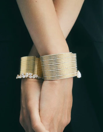 Close-up shot of an adult female's crossed wrists adorned with multiple gold and diamond bracelets, set against a dark green background.