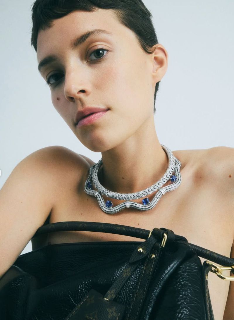 A woman with short dark hair poses with a blue and white jeweled necklace and a dark leather handbag in a fashion shot.