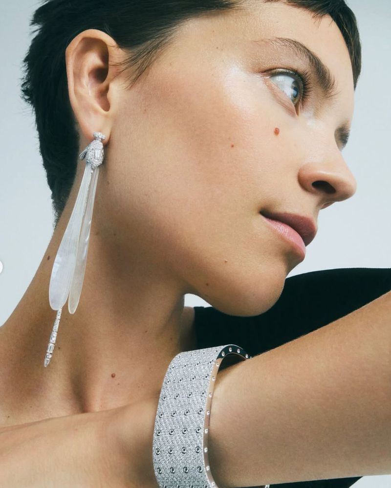 Close-up of a woman with short dark hair, wearing large dragonfly earrings and a jeweled bangle bracelet, against a neutral background.