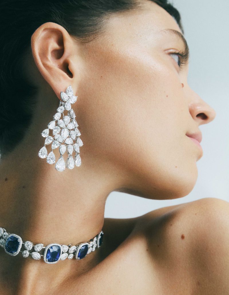Close-up of a woman wearing a diamond chandelier earring and a blue sapphire and diamond choker necklace in a studio setting.