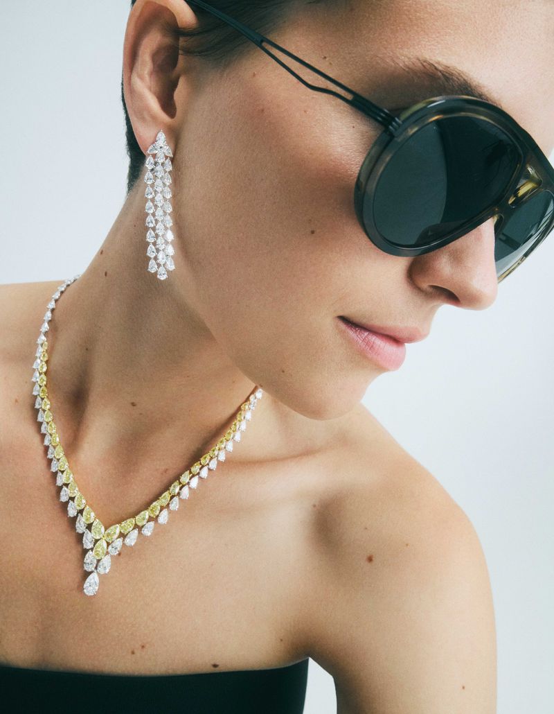 Woman wearing a yellow and white diamond necklace and chandelier earrings with sunglasses against a bright studio background.