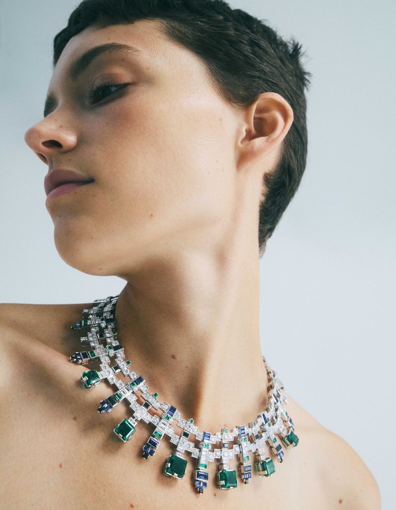 Close-up of a person wearing an elaborate necklace with green, blue, and white gemstones against a plain grey studio backdrop.