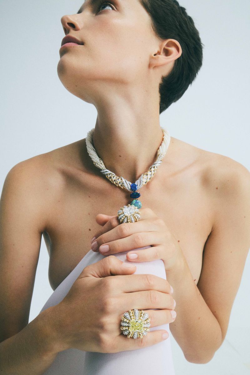Woman poses in a bright studio wearing an ornate beaded necklace with jewel accents, and a large ring with a yellow stone.