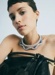 Model with short hair wearing a diamond and sapphire collar necklace holding a black leather bag against a plain white studio backdrop.