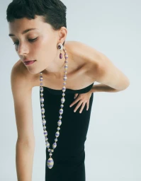 Model with short pixie hair wearing a long purple and gold beaded statement necklace and strapless black dress against a white background.