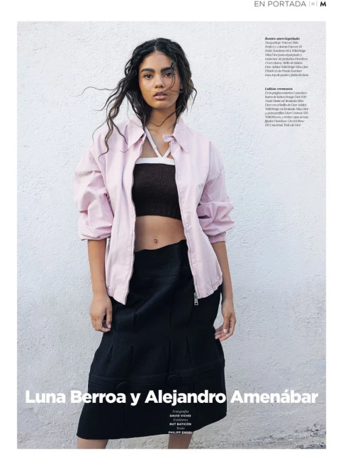 Full-length portrait of a young woman in a pink jacket and black skirt, captured against a textured neutral background in daylight.