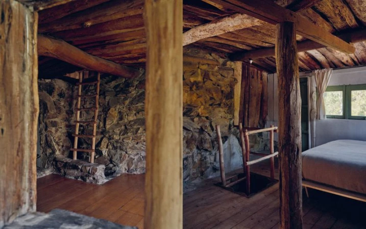 Interior view of rustic cabin features stone walls, exposed wooden beams, and a wooden ladder leading to a loft space.