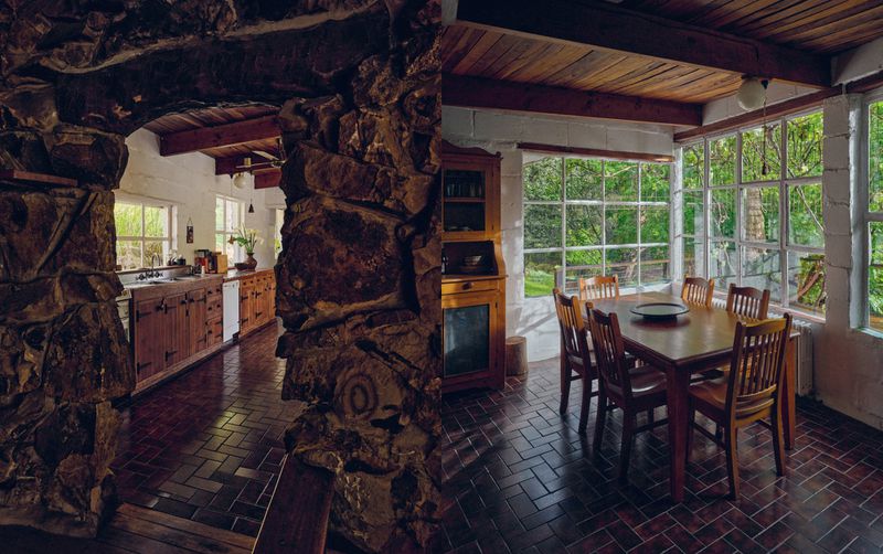 Rustic indoor scene of dining area next to kitchen featuring a wooden table, chairs, stone archway, brick flooring, and natural light.