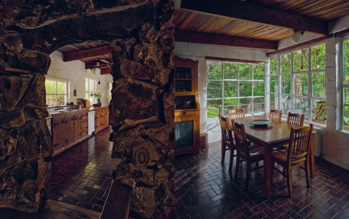 Rustic indoor scene of dining area next to kitchen featuring a wooden table, chairs, stone archway, brick flooring, and natural light.