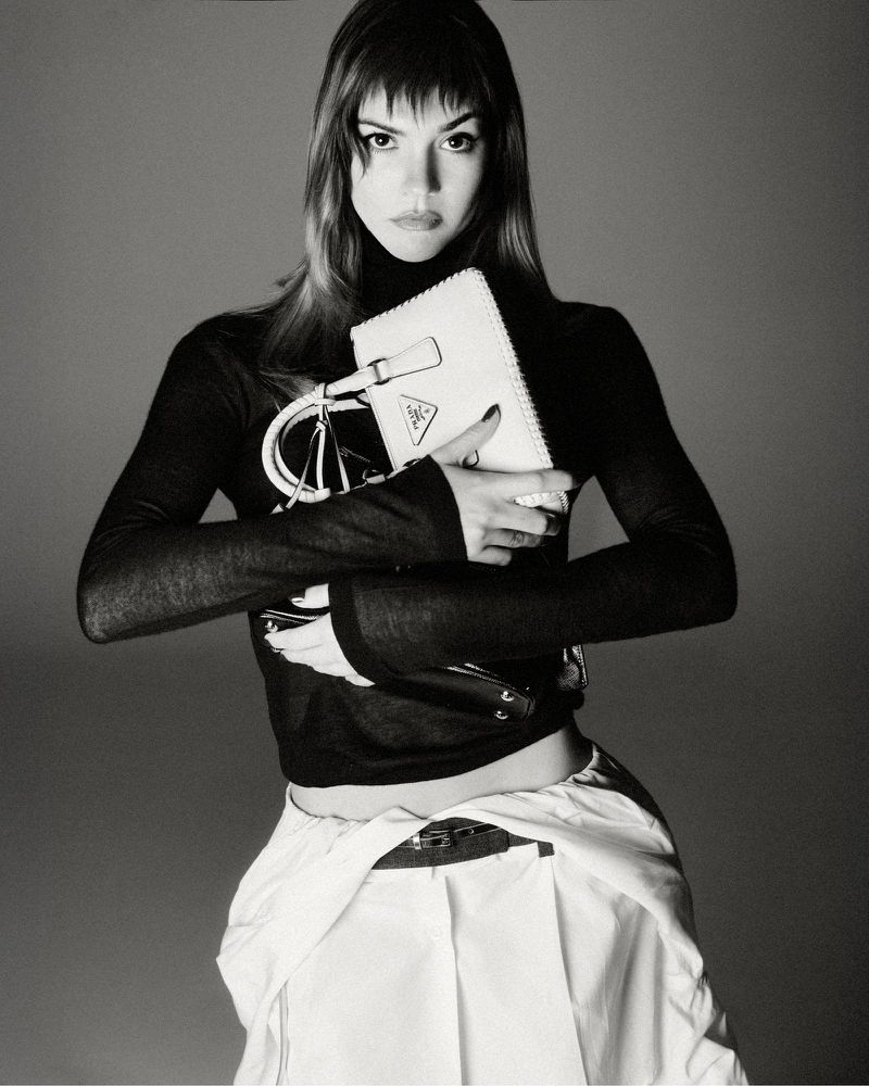 Black and white studio portrait of a woman with short hair holding a white Prada purse, wearing a black shirt and white pants.