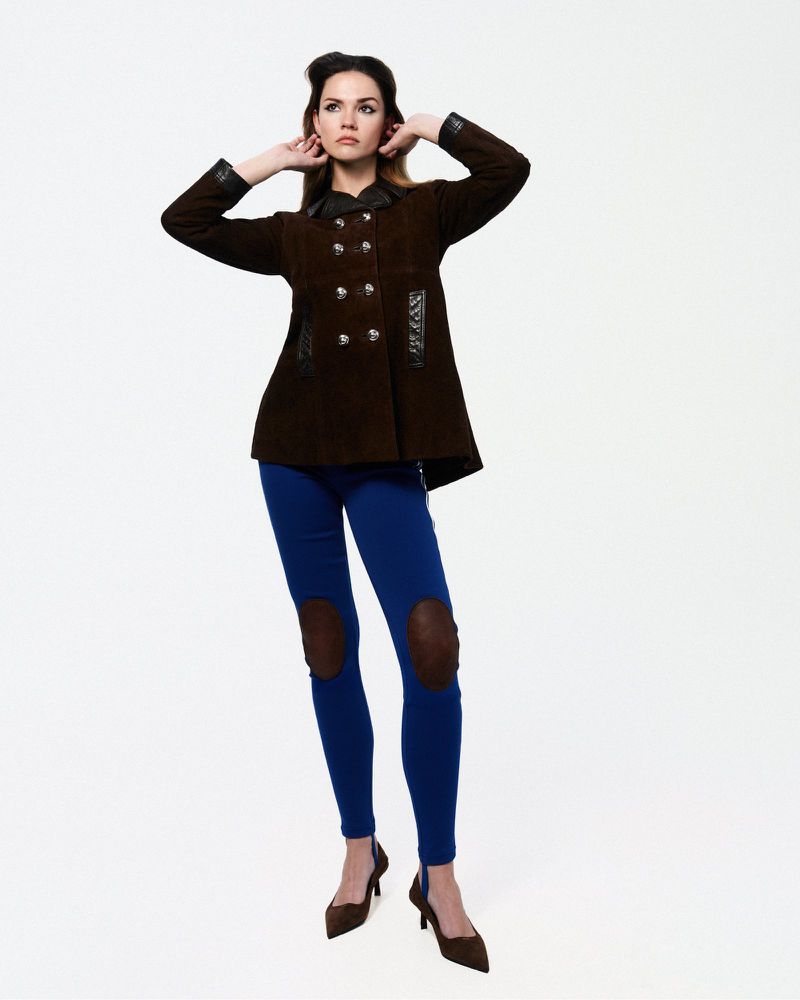 A woman in a vintage-style brown jacket, blue jeans with knee patches, and brown heels strikes a pose against a clean white backdrop.