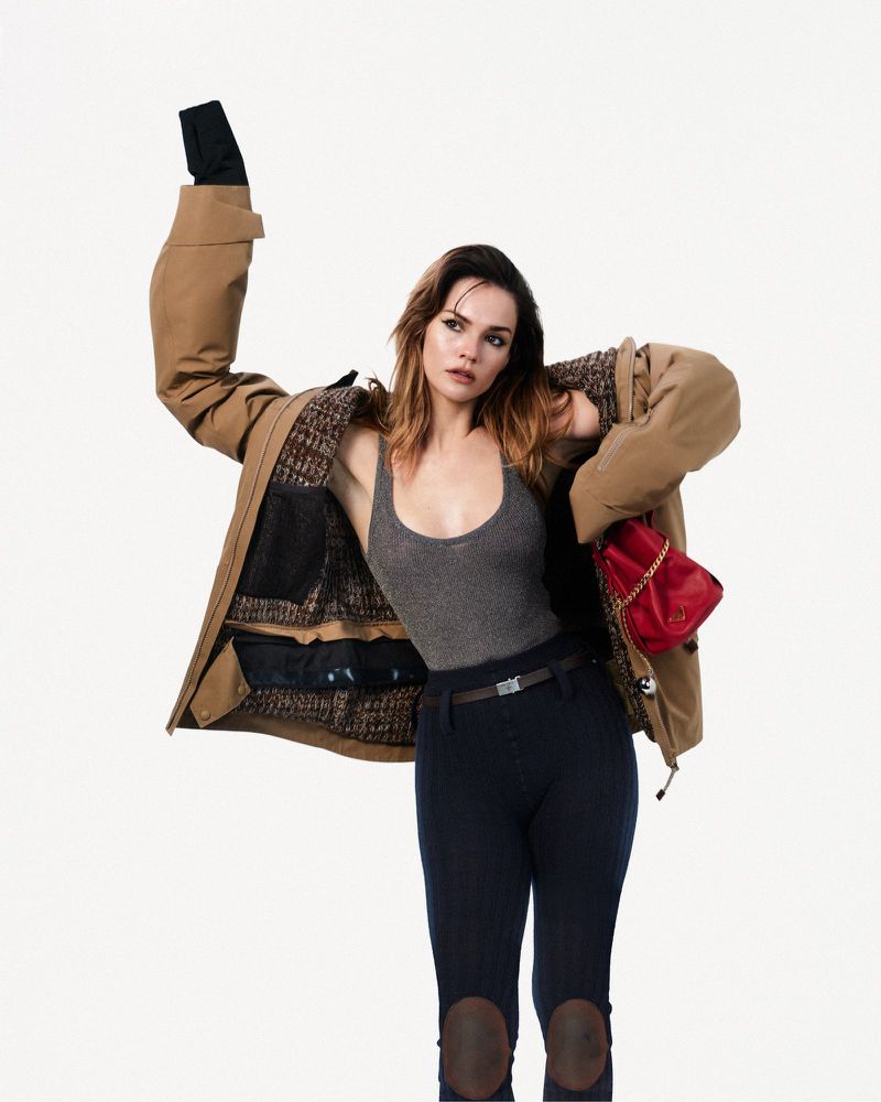 Maia Mitchell wearing an oversized tan jacket, grey tank top, and riding pants against a white background.
