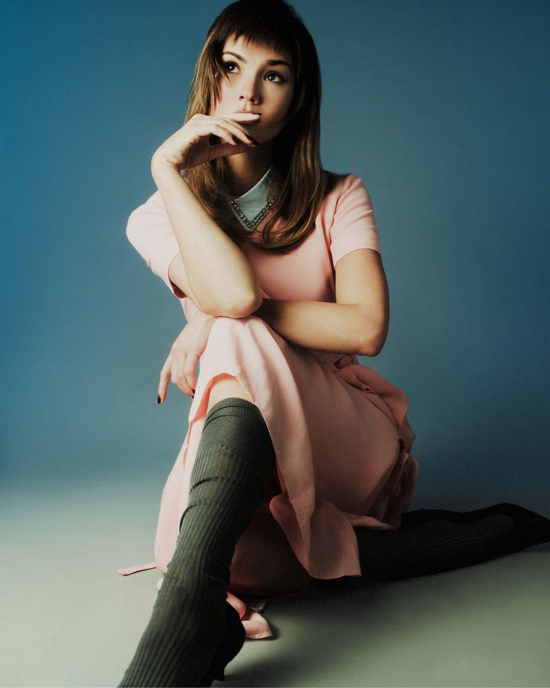 Young woman with bangs wearing a pink dress and knee-high socks, seated against a blue backdrop, posing with a thoughtful expression.