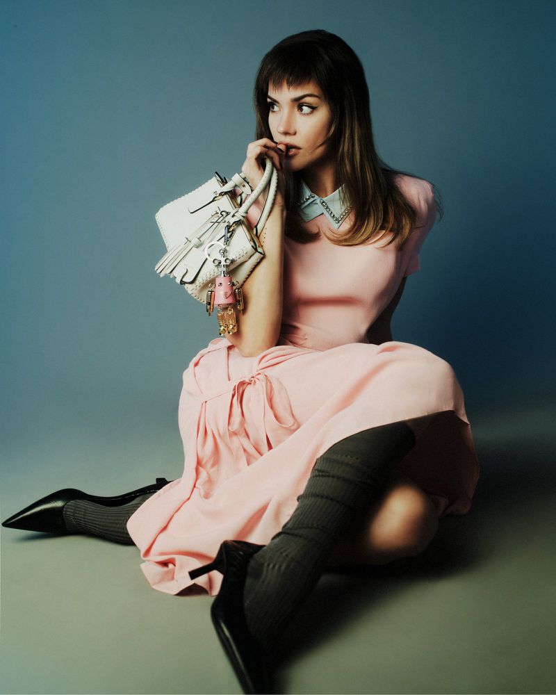 Blonde woman in pink dress sits on the floor, holding a white purse with charms, wearing black heels and grey socks against a blue background.