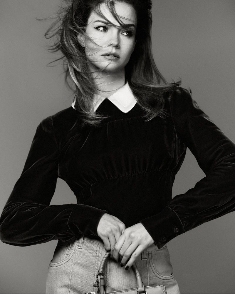 Black and white portrait of a young woman in a long-sleeved top and jeans, holding a handbag and looking to the side.