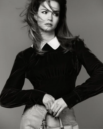 Black and white portrait of Maia Mitchell wearing a black collared velvet blouse and light pants while holding a handbag.