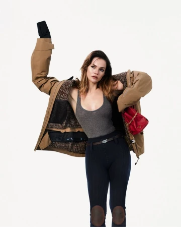 Maia Mitchell wearing an oversized tan jacket, grey tank top, and riding pants against a white background.