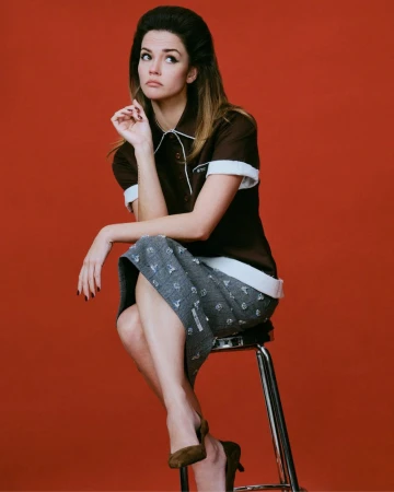 Maia Mitchell wearing a brown collared polo and grey embellished skirt while seated on a stool against a solid red backdrop.
