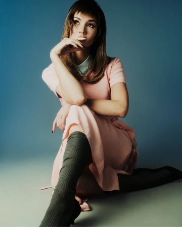 Maia Mitchell seated in a light pink short-sleeve dress and knee-high socks against a blue gradient studio backdrop.