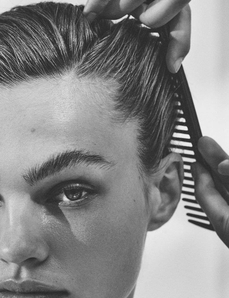 Close-up black and white shot of a young adult's face with short, dark hair being combed by an unseen person, focusing on hair and face.