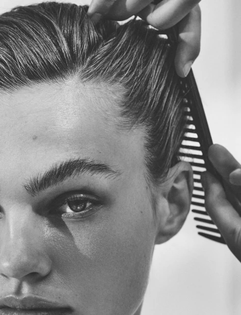 Close-up black and white shot of a young adult's face with short, dark hair being combed by an unseen person, focusing on hair and face.
