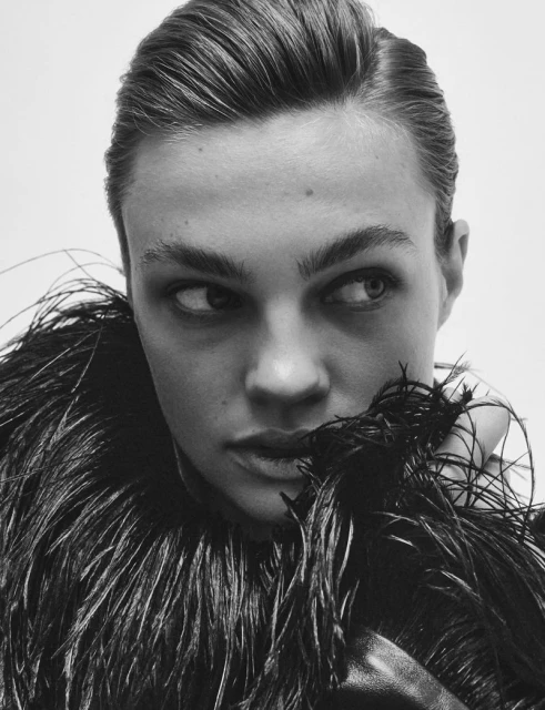 Black and white close-up of a person with sleek hair and a feather boa, looking off to the side against a plain backdrop.