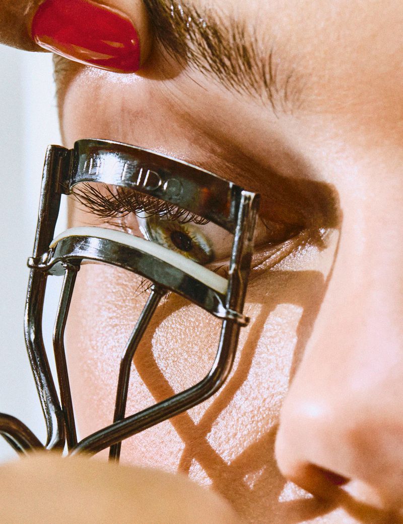 Close-up of a woman using an eyelash curler. The curler is positioned over her eye, with a red fingernail visible in the frame.