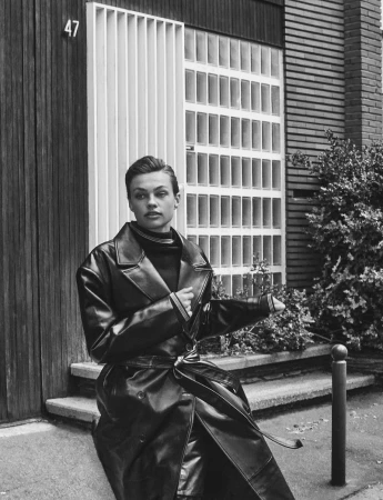 Black and white portrait of a woman in a dark leather coat sitting on steps outside a modern building with geometric details.