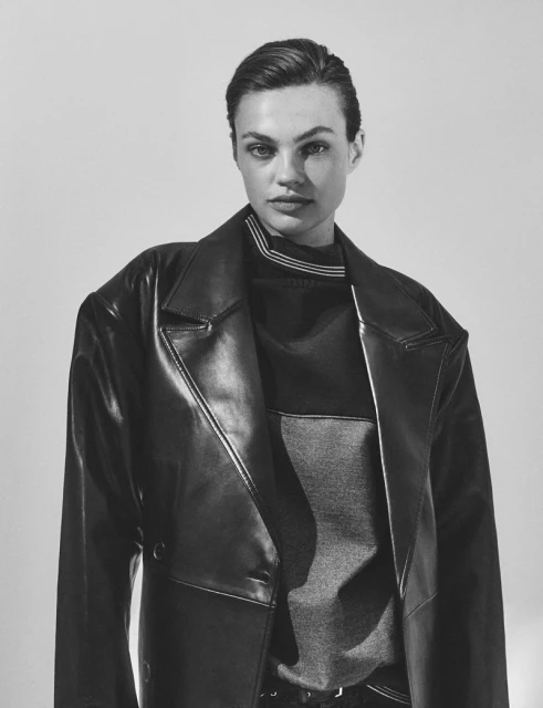 Portrait of a woman with short hair wearing a black leather trench coat in a studio setting. The image is black and white.