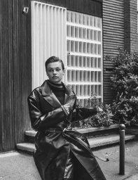 Model Anne Boomgaart wearing a belted black leather trench coat outdoors in front of a glass block facade in black and white.