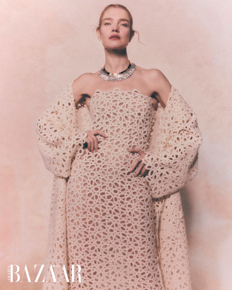 A woman with fair skin poses in a cream-colored, laser-cut gown and jacket, accented by a dark choker necklace, against a neutral backdrop.