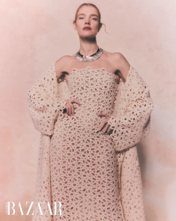 A woman with fair skin poses in a cream-colored, laser-cut gown and jacket, accented by a dark choker necklace, against a neutral backdrop.